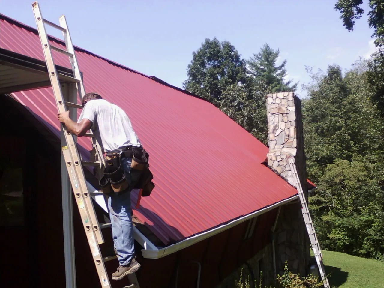 Roofer on a ladder beside a red metal roof and stone chimney — Soffit & Fascia Repair by Roofing Brothers in Windber